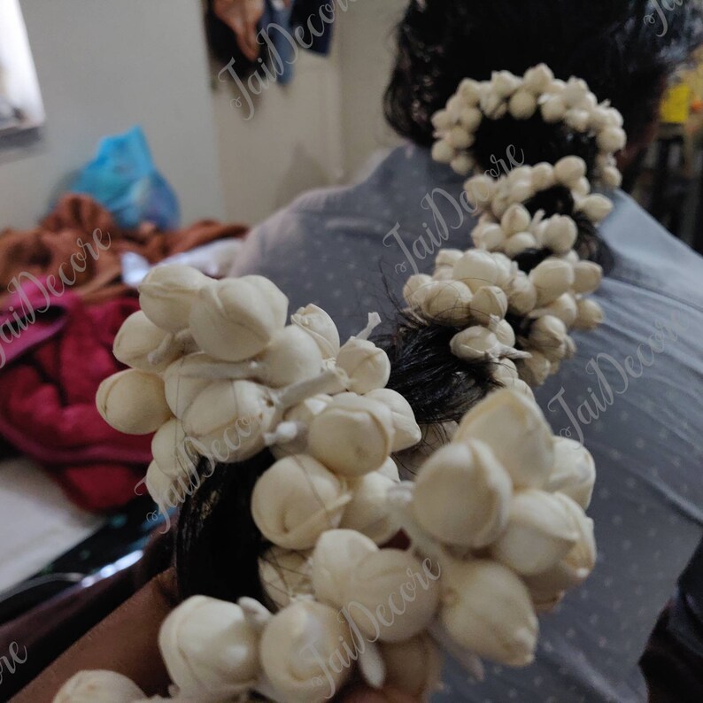 Pu&ograve; includere: Primo piano di una ghirlanda di fiori bianchi, composta da piccoli fiori bianchi rotondi, disposti in fila. La ghirlanda &egrave; legata ai capelli di una persona.