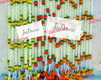 May include: A wall decorated with garlands of white and orange artificial flowers. The garlands are made of small, round flowers and are strung together with green leaves. The garlands are arranged in rows and are hanging from the top of the wall. The text "JaiDecor" is visible on the wall.