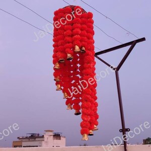 May include: A red and gold decorative hanging garland with pom-poms and bells. The garland is hanging from a metal pole.