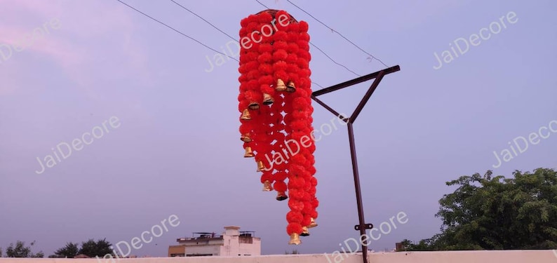 May include: A red and gold decorative hanging garland with pom-poms and bells. The garland is hanging from a metal pole.
