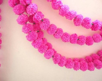 May include: A garland of pink fabric flowers strung together with green thread. The garland is hanging on a light pink wall.