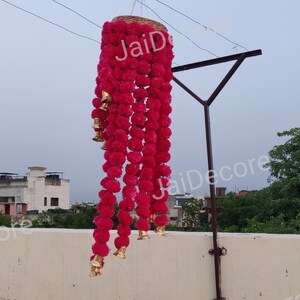 May include: A pink flower garland with gold bells hanging from a ceiling. The garland is made of artificial flowers and is a popular decoration for Indian weddings and festivals.