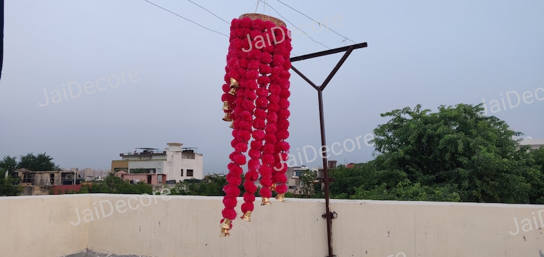 May include: A pink flower garland with gold bells hanging from a ceiling. The garland is made of artificial flowers and is a popular decoration for Indian weddings and festivals.