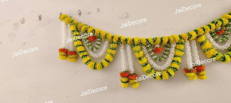 May include: A yellow and green floral garland with white and orange accents. The garland is made of artificial flowers and is hanging on a white wall.