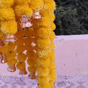 May include: A garland of yellow marigold flowers with gold bells hanging from it. The garland is hanging from a white wall.