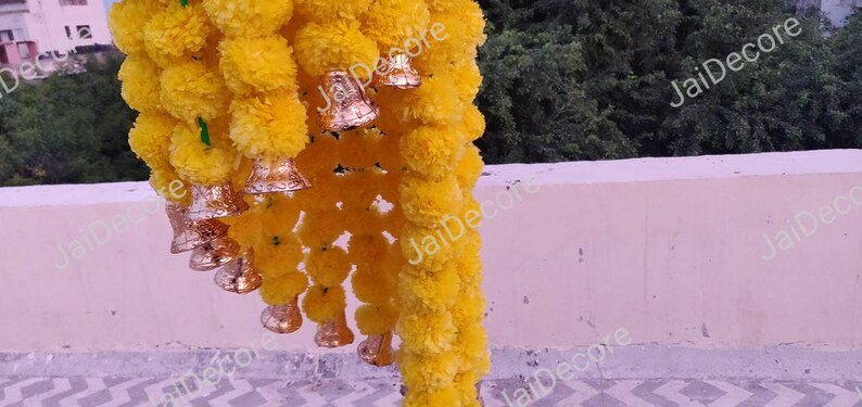 May include: A garland of yellow marigold flowers with gold bells hanging from it. The garland is hanging from a white wall.