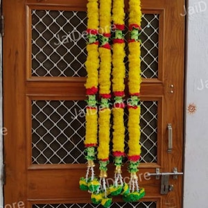Puede incluir: Cinco guirnaldas de flores amarillas con detalles verdes y rojos cuelgan de una puerta de madera. Las guirnaldas están atadas con una cuerda blanca y tienen un borla en la parte inferior.