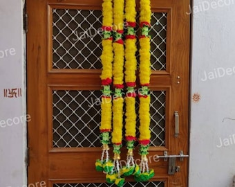 May include: Five yellow flower garlands with green and red accents hang from a wooden door. The garlands are tied together with a white string and have a tassel at the bottom.