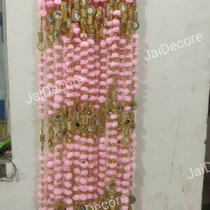 Puede incluir: Un adorno de puerta decorativo rosa y dorado con pequeños pompones y espejos redondos. El adorno se muestra en una percha de madera.