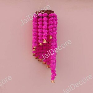 May include: A pink fabric garland with small pom-poms and gold bells hanging from a wooden base. The garland is hanging against a pink wall.