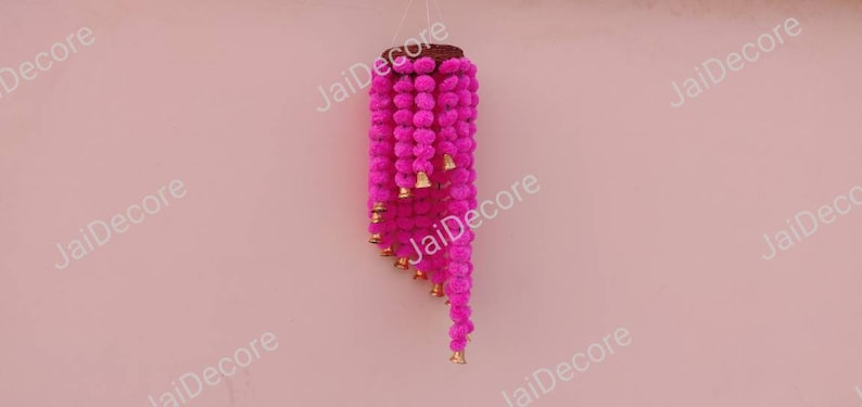 May include: A pink fabric garland with small pom-poms and gold bells hanging from a wooden base. The garland is hanging against a pink wall.