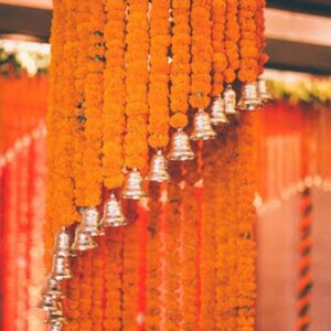 May include: Orange marigold flower garlands with silver bells hanging from a ceiling. The garlands are arranged in a cascading pattern.