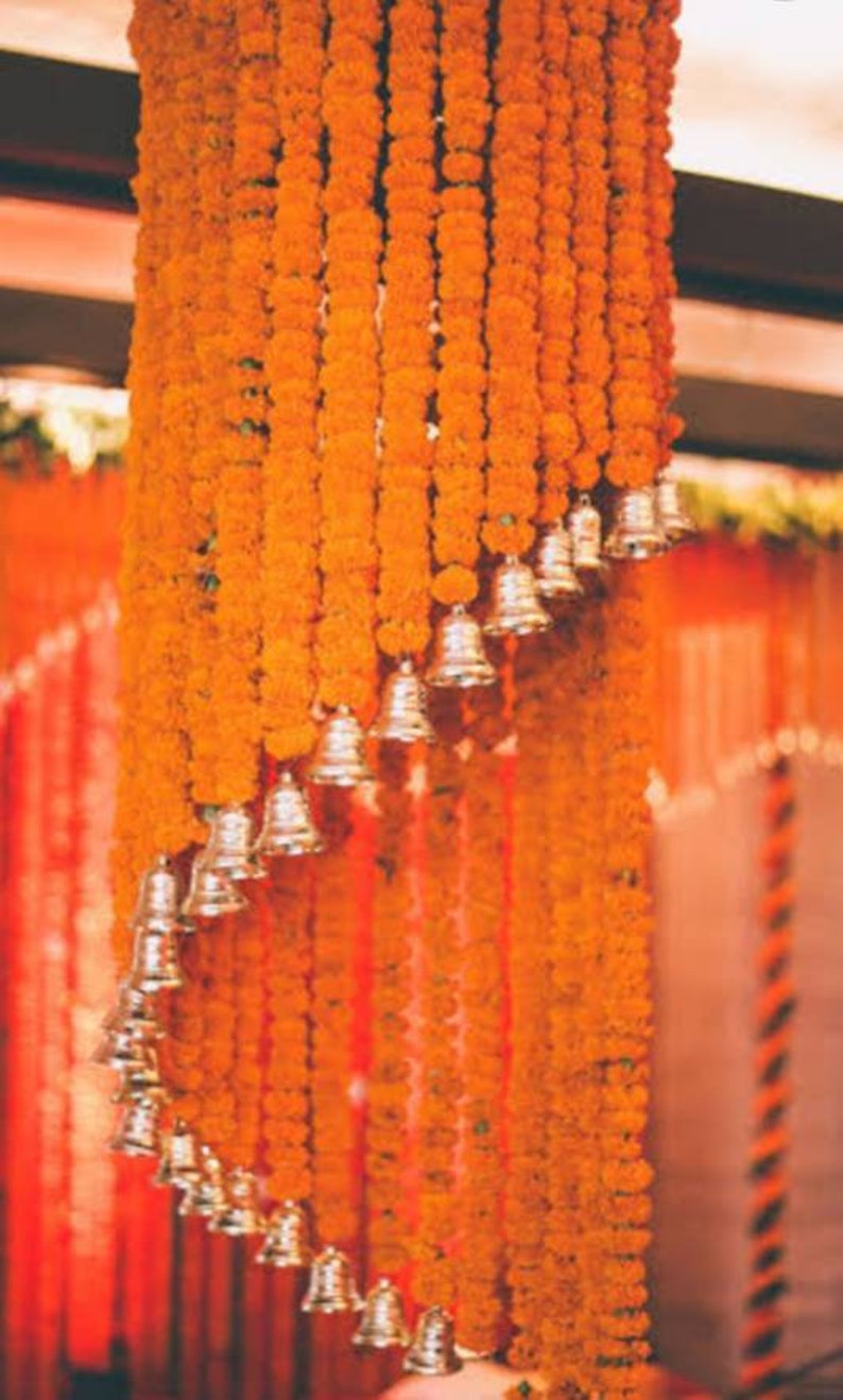 May include: Orange marigold flower garlands with silver bells hanging from a ceiling. The garlands are arranged in a cascading pattern.
