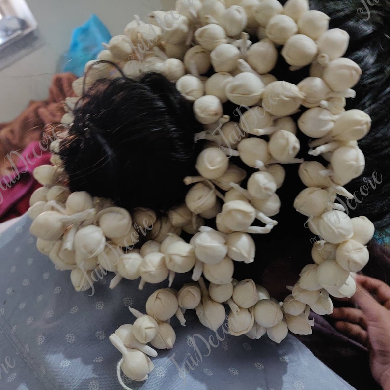 Pu&ograve; includere: Una ghirlanda di fiori bianchi fatta di piccoli fiori rotondi di stoffa bianca. La ghirlanda &egrave; avvolta intorno ai capelli di una persona.