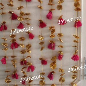 May include: A set of five gold and pink decorative garlands with tassels. Each garland features gold paper circles and pink tassels. The garlands are hanging from a white wall. The text "JaiDecor" is visible on the garlands.