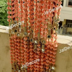 Puede incluir: Una cadena de pompones de color melocotón con detalles dorados y pequeños espejos redondos. La cadena está colgada de una barandilla y tiene el texto "JaiDecor" impreso.