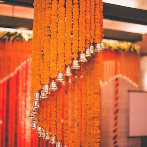 May include: A wedding ceremony decoration made of orange marigold flower garlands with silver bells hanging from them.