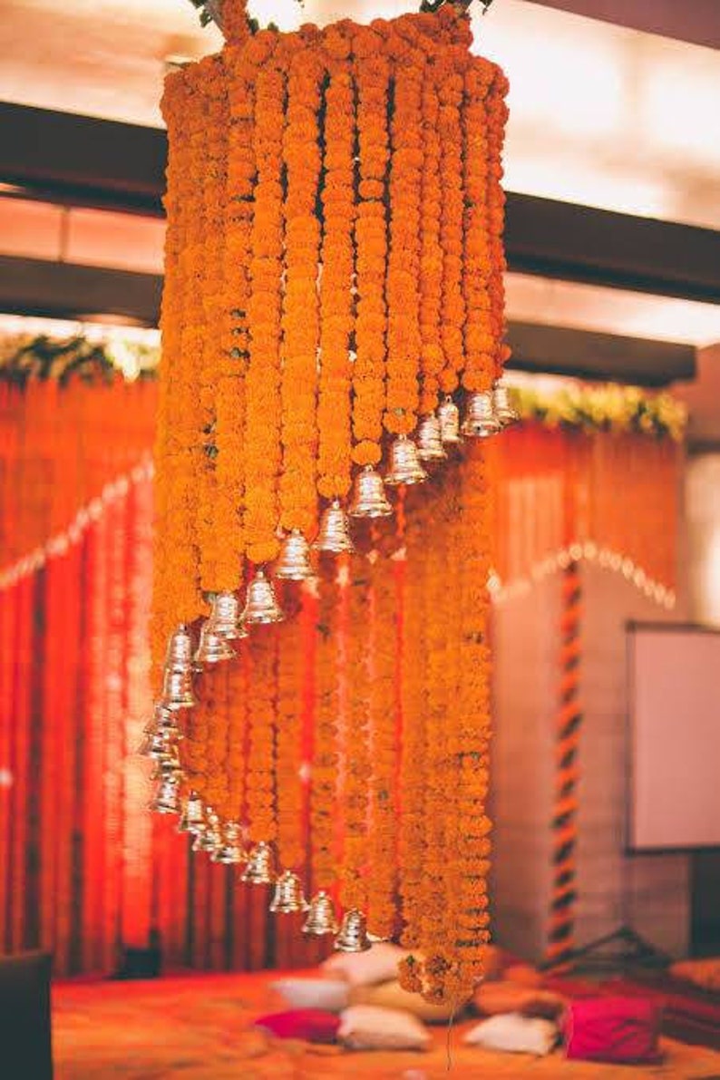 May include: A wedding ceremony decoration made of orange marigold flower garlands with silver bells hanging from them.