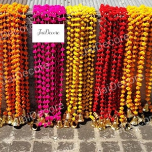 Puede incluir: Cinco guirnaldas decorativas con pompones en naranja, rosa, amarillo y rojo. Cada guirnalda tiene cascabeles dorados en la parte inferior. El texto "JaiDecor" es visible en una etiqueta blanca.