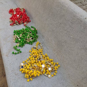 Puede incluir: Tres hilos de guirnaldas decorativas de cuentas en rojo, verde y amarillo. Cada hilo presenta pequeños pompones redondos y coloridos y detalles decorativos espejados. Las guirnaldas están dispuestas sobre una superficie de hormigón gris.