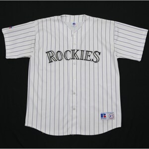 May include: A white short-sleeved baseball jersey with thin vertical navy blue pinstripes. The word ROCKIES is embroidered in black and silver across the chest. A Russell Athletic brand tag is visible on the lower front and the sleeve.