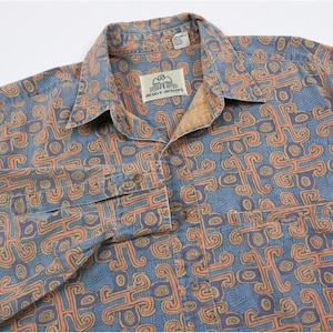 May include: A button-down shirt with a blue background and a repeating pattern in orange and purple. The shirt has a collar, a chest pocket, and the text "desert designs" on a tag.