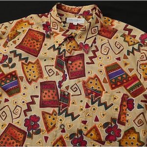 May include: Tan button-up shirt with a colorful geometric pattern. The shirt features a variety of shapes, including squares, triangles, and zigzags, in shades of red, orange, yellow, and purple. The shirt has a collared neckline and a pocket.