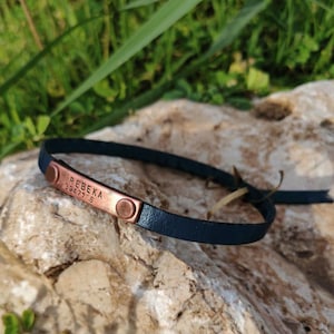 Peut inclure: Un bracelet en cuir bleu foncé doté d'une plaque métallique rectangulaire couleur cuivre fixée par deux rivets. La plaque est gravée avec le texte REBEKA et les chiffres 39477 6. Le bracelet présente une fine lanière avec un nœud simple.