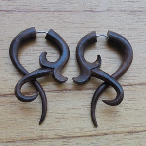 May include: A pair of dark brown wooden earrings with a unique, intricate design. The earrings are shaped like a swirl with a small loop at the top for hanging.