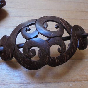 May include: A dark brown coconut shell hair stick with a carved design of three swirls. The stick is about 6 inches long and has a smooth, polished finish.