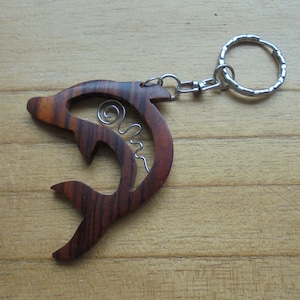 Puede incluir: Un llavero de madera con forma de delfín con un anillo y una cadena de metal plateado. El delfín está tallado en madera oscura con un patrón de veta marrón claro. Un alambre plateado está enrollado dentro del cuerpo del delfín.