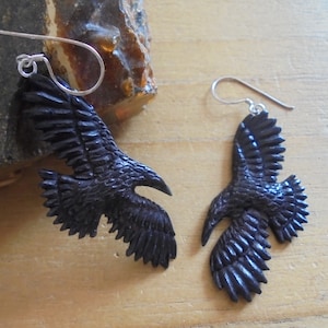 Puede incluir: Un par de pendientes de cuervo negro con ganchos de plata. Los cuervos están tallados en un estilo detallado y realista con las alas extendidas.