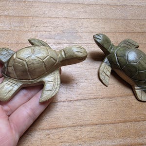 Op de afbeelding: Twee houten schildpadbeeldjes. Eén wordt in de hand gehouden en heeft een lichtbruine kleur. De andere schildpad is donkerder groen. Beide schildpadden hebben gesneden details, waaronder de schild en vinnen. De schildpadden zijn decoratieve items.