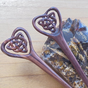 Op de afbeelding: Twee houten haarstokken met hartvormige toppen met ingewikkelde Keltische knoopontwerpen. De stokken zijn donkerbruin met een roodachtige tint.