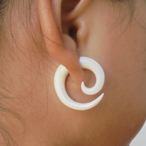 May include: White spiral ear gauge made of bone. The gauge is worn in the earlobe of a person.