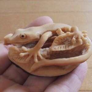 Op de afbeelding: Een klein, ingewikkeld gesneden beeldje van een hagedis of gekko, gemaakt van een lichtbruin materiaal, mogelijk bot of ivoor. De hagedis is afgebeeld met zijn poten uitgestrekt en zijn staart om zijn lichaam gekruld. Het beeldje staat op een voet met een gesneden bladontwerp.