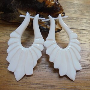Puede incluir: Un par de pendientes de hueso blanco con un diseño tallado en forma de abanico. Los pendientes tienen forma de lágrima con un gran bucle abierto.