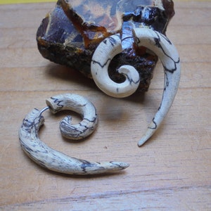 May include: Two wooden spiral ear gauges, one with a simple spiral design and the other with a more intricate spiral design. The gauges are made of light brown wood with dark brown streaks.