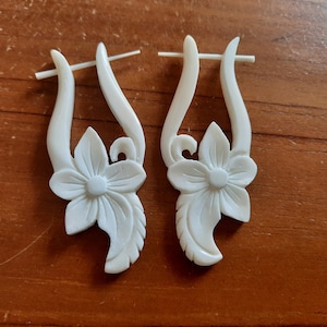 May include: A pair of white carved bone earrings with a floral design. The earrings are shaped like a flower with a curved stem.