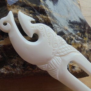 May include: White bone hair fork with intricate carvings in the shape of a fish hook. The fork is resting on a brown and white rock.