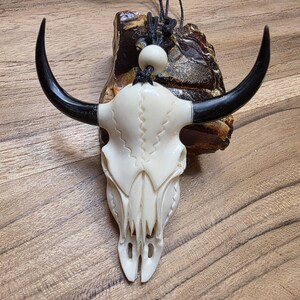 May include: A carved, off-white bull skull pendant with black horns. The skull is detailed with carved lines and attached to a black cord. The pendant is displayed on a piece of rough, brown material, set against a wooden background.