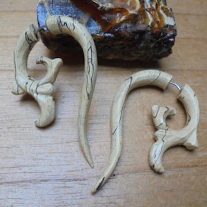 May include: Three wooden earrings, two are curved and one is a more intricate design. The earrings are a light brown color with dark brown streaks.