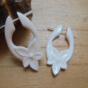 Puede incluir: Un par de pendientes blancos tallados. Cada pendiente presenta un diseño floral en la parte inferior y una forma curva y abierta. Los pendientes están hechos de un material liso y brillante y se exhiben sobre una superficie de madera.