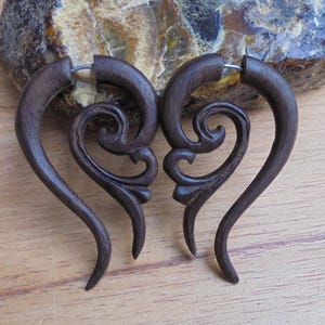 May include: A pair of dark brown wooden earrings with intricate carved designs in the shape of hearts. The earrings are made of wood and have a silver metal hoop.