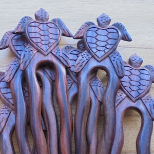 May include: A set of six wooden hair sticks with a carved turtle design. Each stick features a turtle with wings and a heart-shaped shell. The sticks are a dark brown color.