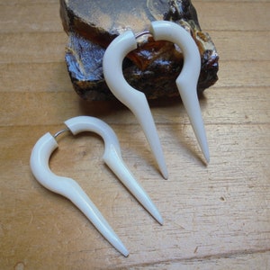 Op de afbeelding: Drie witte plastic oorringen met een puntige punt aan elke kant. De oorringen liggen op een houten oppervlak.