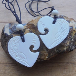 May include: Two white bone heart-shaped pendants with intricate carvings, hanging on black cords with white beads. The pendants are designed to fit together.