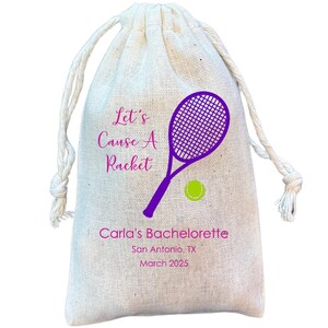 May include: A white cotton drawstring bag with the text "Let's Cause A Racket" in pink and a purple tennis racket and green tennis ball graphic. The text "Carla's Bachelorette" is printed below the graphic with the date and location in smaller font.