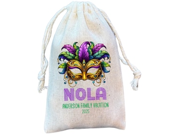 May include: A beige drawstring bag featuring a colorful Mardi Gras mask design. The mask is gold, purple, green, and blue. The bag has the word "NOLA" in purple, with "Anderson Family Vacation 2025" printed below.