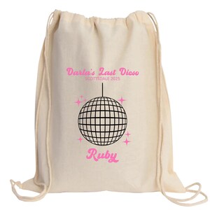 May include: A white drawstring bag with a pink and black design. The bag features a disco ball, stars, and the text "Darla's Last Disco Scottsdale 2023" and "Ruby".